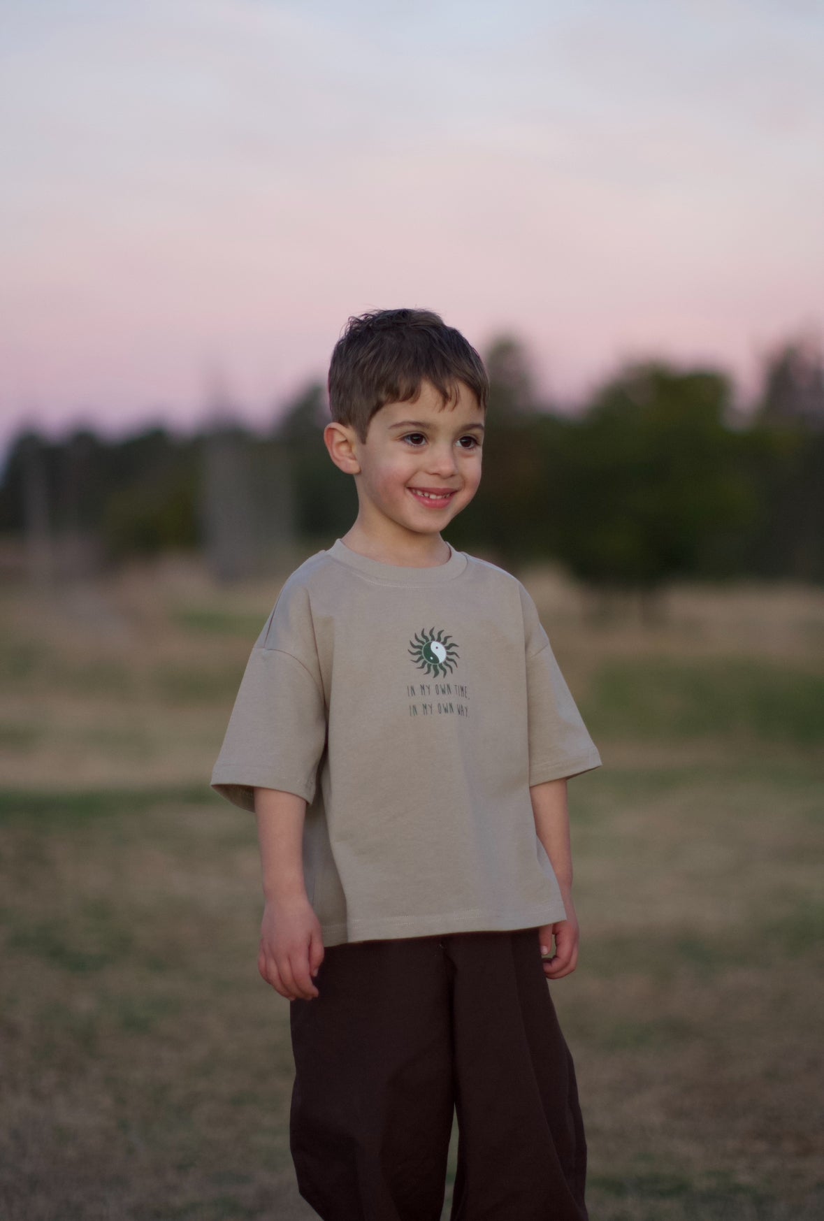 Child wearing beige tee with a chest placement print of a yin and yang sun with text below saying " in my own time, in my own way". 