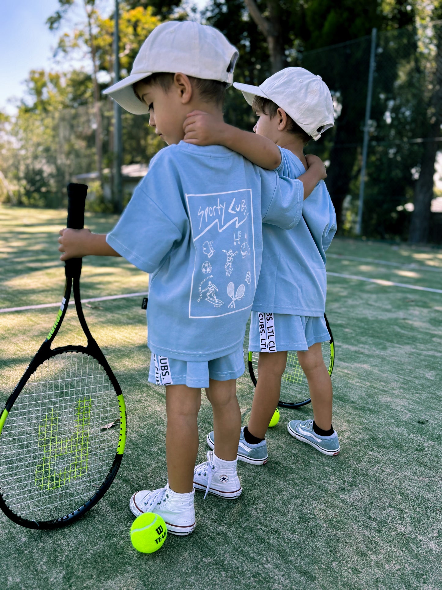 Twin brothers wearing dusty blue sporty kids sets. Posing on the tennis court. Wearing LTL cubs hats. 